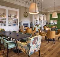 Spacious dining area with wooden floor, featuring a mix of green and brown chairs, tables, and large hanging lamps. Decorative shelves and a green accent wall complete the room's elegant design.