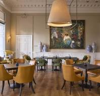 A well-lit, elegant room with tan chairs around tables, a large hanging lamp, and a painting on the wall.