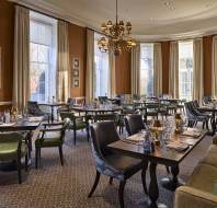 Elegant dining room with neatly arranged tables and chairs. Large windows with curtains and a chandelier overhead provide soft lighting. Neutral tones with green and brown accents throughout.