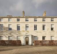 A large, three-story, historic stone building with symmetrical windows and a central entrance. A hedge surrounds the base, and bare trees are visible in the background.