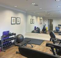 A gym room with exercise equipment including dumbbells, a yoga ball, treadmill, bench, and exercise mat. Mirrors and framed pictures adorn the walls.