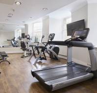Gym with exercise equipment, including a treadmill, stationary bikes, and elliptical machine on wooden flooring. Mirrors reflect the equipment, and a wall-mounted TV is above the radiator.