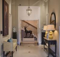 Elegant hallway with a deer statue, art piece, and a staircase. A table with lamps is on the right, a yellow bench on the left, and decorative blue vases are present. Chandelier overhead.