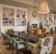 Elegant dining room with a long dark table, eight mixed chairs, and built-in shelves displaying plates and vases. There is a fireplace, a large mirror, and modern lighting overhead.