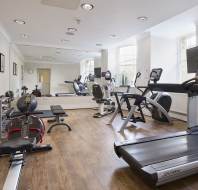 A bright gym room with wooden floors, containing treadmills, exercise bikes, a weight bench, and mirrors. Large windows let in natural light.