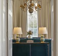 Elegant room with a chandelier, two lamps on a table, a large window with curtains, and a decorative bouquet. A velvet sofa is partially visible in front of the table.