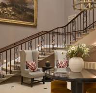 Hotel lobby featuring a staircase, two armchairs with patterned cushions, a large vase with flowers on a glossy round table, and a painting of sheep on the wall.