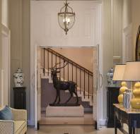 Elegant hallway with a stag sculpture in front of stairs, a sofa, blue vases on black tables, round mirror, lamps, and a framed painting.