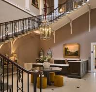 Elegant hotel lobby with a grand staircase, chandelier, art on walls, round table with flowers, and seating area. Neutral tones dominate the decor. A doorway leads to a room labeled "The Morning Room.