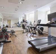 A gym with treadmills, stationary bikes, a rowing machine, free weights, and benches on a wooden floor. Mirrors and a wall-mounted TV are also visible.
