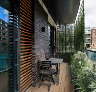 Modern balcony with a wooden table and two chairs, surrounded by greenery. Slatted wood panels and glass railings contrast the brick walls and nearby buildings.