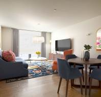 Modern living room with a round wooden dining table, blue and orange chairs, a navy sofa, floral rug, large TV, wall art, and a window with light gray curtains.