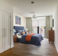 Bedroom with a large bed featuring blue and orange bedding, two bedside tables with lamps, a dresser, and a large window. Walls are white, with a patterned accent wall behind the bed.