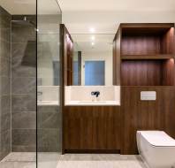 Modern bathroom with gray tiled shower, wooden vanity with sink and mirror, and toilet. Shelving unit incorporated next to the toilet.
