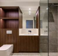 Modern bathroom with brown wooden cabinetry, a white toilet, and a glass-enclosed shower with grey tiles. A wall-mounted mirror with built-in lights is above the sink.