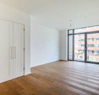 Empty room with wooden floors, white walls, two closed white doors on the left, and a large window with blinds overlooking an exterior view of buildings.