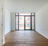 Empty room with wooden flooring, white walls, and a large window facing outside. A door is visible on the left.