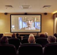 People in a dimly lit room watch a film on a large screen, featuring a woman in period costume.