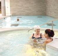Two people relax in a jacuzzi talking, while another person swims in a larger pool in a spa setting.