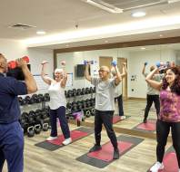 A fitness instructor guides three people lifting dumbbells in a gym mirrored room.