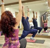 Four people in workout clothes perform yoga poses on mats in a gym with mirrored walls and gym equipment in the background.