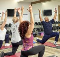 A group of four people practice yoga in a fitness room, kneeling on mats with arms raised. Mirrors and exercise equipment are visible in the background.