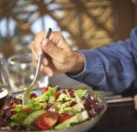 Person in a blue shirt eating a salad with a fork, next to a glass of red wine on a table.