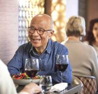 Older adults dining at a restaurant, engaging in conversation. Two glasses of red wine and a salad are on the table.