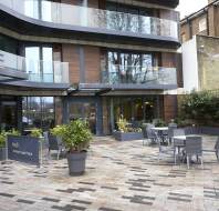 Outdoor seating area of a modern building with glass balconies and planters. Signs read "No3 at Nightingale Place." Tables and chairs are arranged on a patterned paved surface.