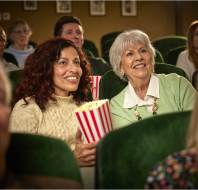 Two women sitting in a theater, smiling and holding popcorn, surrounded by other people seated in rows.