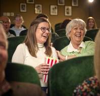 Audience seated in a cinema, with a young woman holding popcorn and an older woman beside her, both smiling and laughing.