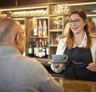 Barista in an apron serves a cup to a seated customer at a counter. Shelves with bottles and a coffee machine are in the background.
