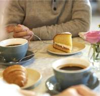 Two people sitting at a table with coffee cups, a slice of cake, and a croissant. A vase with pink and purple flowers is on the table.