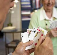 Two people playing cards at a table. One person holds a hand with an ace of clubs, queen of hearts, five of spades, king of hearts, and four of hearts. The other person smiles, holding their cards.