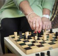 Person in a green top playing chess, moving a piece on a wooden chessboard with both black and natural-colored pieces. They are seated and wearing a bracelet and watch.