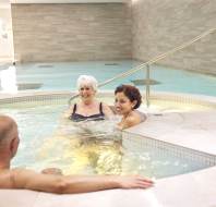 Three people relaxing in a hot tub at an indoor pool facility. Two women are seated in the water, facing a man who's sitting on the tub's edge.