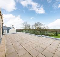 Spacious rooftop terrace with beige tiles and glass railing overlooking a countryside landscape under a partly cloudy sky.