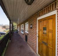 View of a brick exterior hallway with a wooden door labeled '36' on the right, windows on the left, and parked cars outside a fence in the distance.