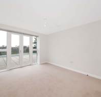 An empty, carpeted room with neutral walls, glass doors leading to an outdoor area, and a view of greenery outside.