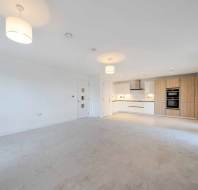 Spacious, empty open-plan living area with beige carpet and white walls. Kitchen has light wood cabinetry and modern appliances. Large windows allow ample natural light.
