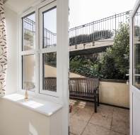 A room with patterned curtains, a glass vase on the windowsill, and an open door leading to a small outdoor patio with a wooden bench and plants.