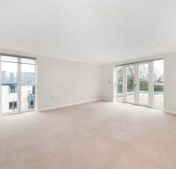 Spacious, empty room with beige carpet, white walls, and ceiling. Features large windows and glass doors leading to a balcony with a view of neighboring buildings and trees.