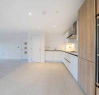 Modern open-plan kitchen with white cabinets, wood-paneled appliances, and tile flooring, adjacent to a carpeted living area. Bright lighting.