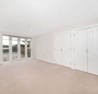 Empty room with beige carpet, white walls, two sets of double doors, and a large window leading to a patio with a view of greenery.