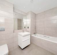 Modern bathroom with beige tile walls and floor, featuring a bathtub, wall-mounted toilet, sink with cabinet, large mirror, and recessed ceiling lights.