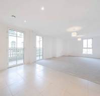 Bright, empty living room with white walls, tiled and carpeted flooring, two sets of windows, and ceiling lights. Large glass double doors open to a balcony.