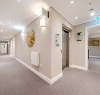 A brightly lit hallway with beige walls and grey carpet, featuring an elevator, wall decorations, framed pictures, and a visible exit sign.