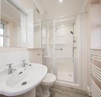 Bathroom with a white sink, dual taps, and mirror, adjacent to a shower with sliding doors. Walls are light-colored with a heated towel rail and frosted windows.