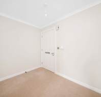 An empty room with beige carpet, white walls, and a white door. A single light bulb hangs from the ceiling.