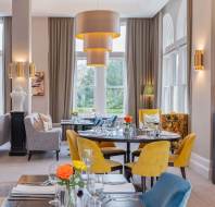 Elegant restaurant dining area with tables set for service, yellow and blue chairs, large pendant light, and large windows with drapes.
